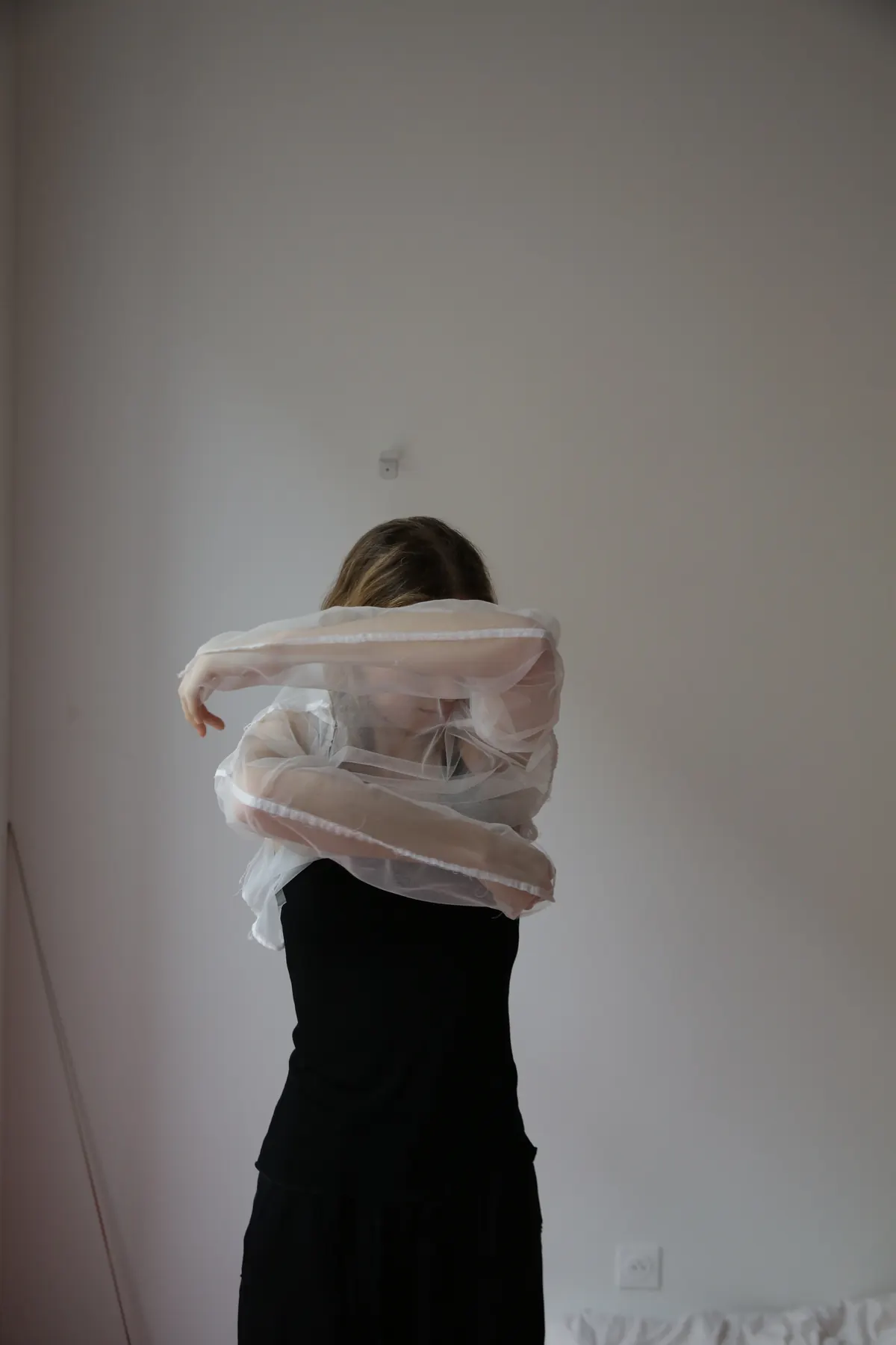 Model wearing a sheer white organza blouse over a black dress, arms folded across her face against a pale wall